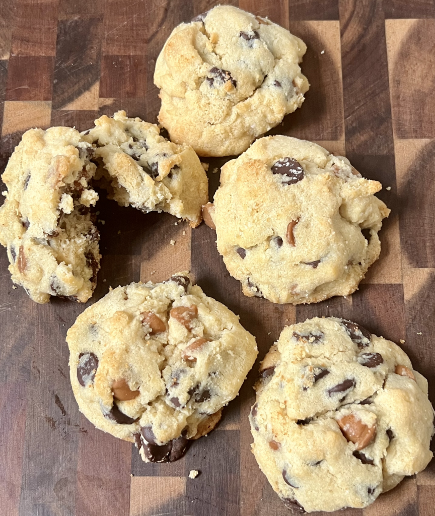 Keto peanut butter chocolate chip cookies stacked on a wooden board with one cookie broken open to show the soft, gooey center